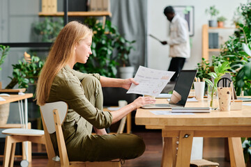 Side view of young businesswoman with long blond hair analyzing financial data in document and on screen of laptop