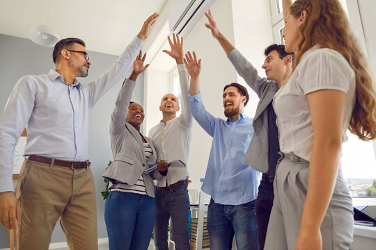 Positive motivated colleagues raise their hands together symbolizing their goal for success. Happy men and women in office are getting ready for work. Concept of unity and teamwork. Angled bottom view