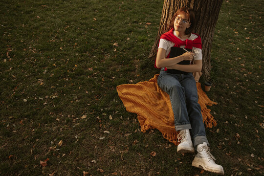 Relaxed young caucasian woman sits on grass by tree with eyes closed and drawing pad close to her. Brown-haired woman in casual autumn clothes. Modern education, student body concept.