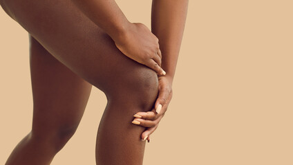 Young black lady whose knee is hurting is holding her hand on injured knee cap area. Cropped shot of human legs isolated on beige color background. Injury, rheumatoid arthritis, feeling pain concept