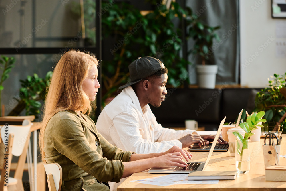Poster side view of two young intercultural designers or analysts networking by desk in front of laptop scr