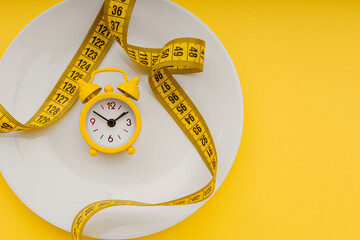 Yellow alarm clock on a white plate with measuring tape on a yellow background. Top view