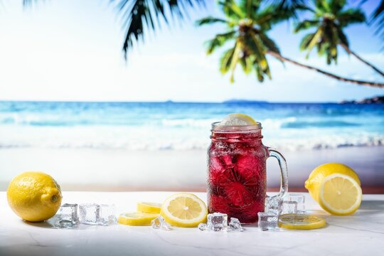 Closeup Of A Red Summer Drink In Transparent Glass And Yellow Lemons With A Topical Beach Background