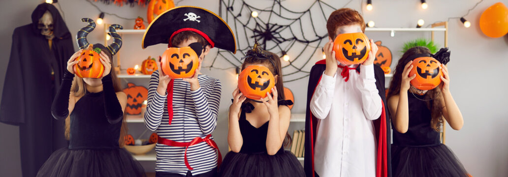 Narrow web banner shot of cute small children dressed in costumes for Halloween celebration have fun in kindergarten together. Excited kids with jack-o pumpkins enjoy all saints eve party or carnival.