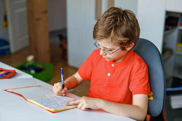Naklejka na ściany i meble Portrait of little kid boy with glasses at home making homework, writing and learning. Little child doing exercise, indoors. Elementary school and education, home schooling concept.