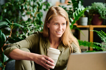 Young smiling businesswoman looking at laptop screen while communicating in video chat and having coffee by workplace