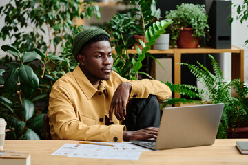 Young serious black man in casualwear sitting by workplace in front of laptop screen in green...