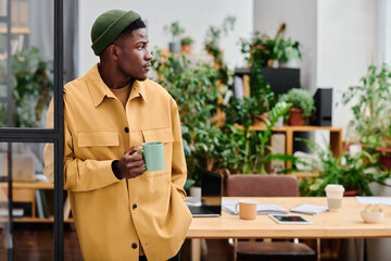 Young black man in stylish casualwear holding cup of coffee while standing in front of camera...