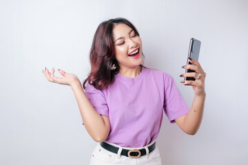 A portrait of a happy Asian woman is smiling and holding her smartphone wearing a lilac purple t-shirt isolated by a white background