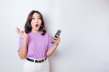 Surprised Asian woman wearing lilac purple t-shirt pointing at her smartphone, isolated by white background