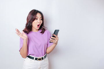 Fototapeta premium Surprised Asian woman wearing lilac purple t-shirt pointing at her smartphone, isolated by white background
