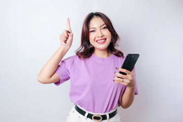 Smiling beautiful Asian woman pointing finger upward to empty space while holding her phone isolated white background