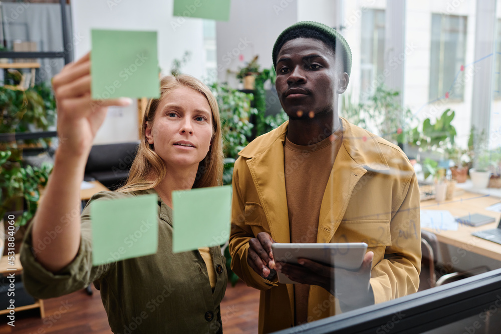 Poster young confident businesswoman pointing at notepaper with working tasks while standing next to collea