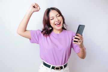Naklejka premium A young Asian woman with a happy successful expression wearing lilac purple shirt and holding smartphone isolated by white background