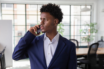 Serious African-American employee talking on phone in office. Medium shot of pensive young male office worker standing, looking away, negotiating with business partner. Business, communication concept