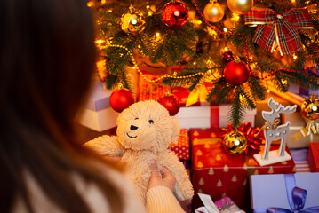 Back view of a girl holding teddy bear present on background of Christmas tree. Present for winter holidays concept, warm home atmosphere