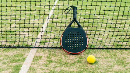 Yellow ball on grass turf near padel tennis racket behind net in green court
