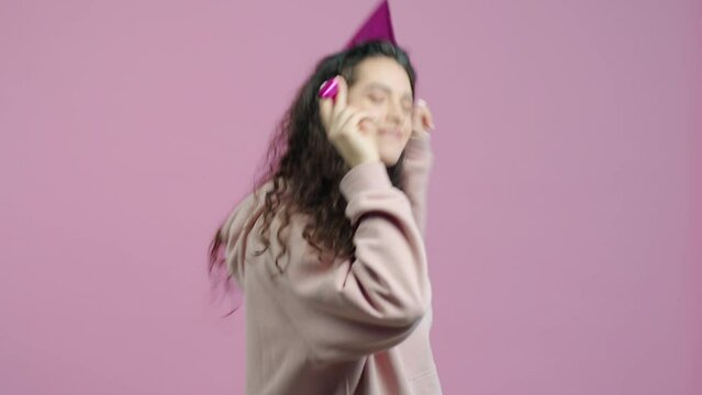 Portrait of excited young lady wearing party hat dancing and blowing horn having fun against pink background. Celebrations and joy concept.