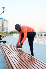 Side view of young African American man in activewear tying shoelace on sneaker while bending over leg bent in knee in the street