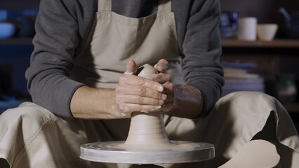 An unrecognizable potter sculpts soft clay with wet hands on a potter's wheel. Clay shaping and sculpting, close up hand creative work in a working workshop. Processing pottery material.