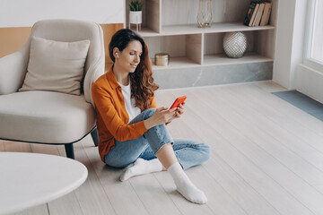 Girl in earphones using phone app, listen to music, audio book or podcast, sitting on floor at home