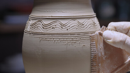 Male hands of a potter carve designs on a soft white clay pot with pottery tools. A man decorates a pot with a serrated scraper, drawing lines and waves. Unfired earthenware in a workshop close up.