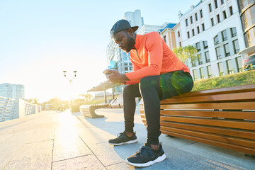 Young tired African American athlete in activewear texting in smartphone while sitting on bench in the morning and having rest after training