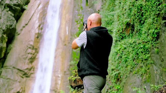 male photographer photographing waterfall giessbach in alpine mountains, fast flowing powerful stream of clear water, splashes scatter, concept of wild nature, natural resources, active sports tourism