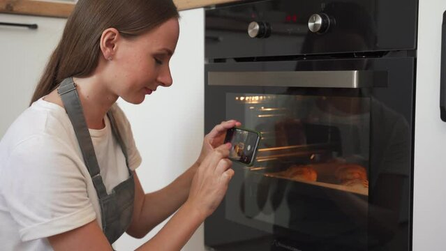 Charming young woman in a grey chef's apron puts cookies in the oven and takes pictures on her phone. 