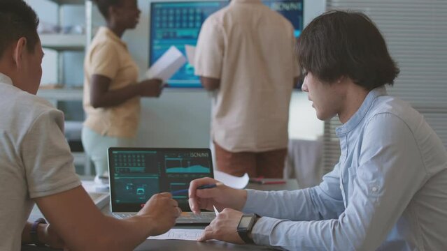 Two Asian male engineering students looking at environmental indexes on laptop screen and discussing project on renewable energy and sustainability, sitting at office desk with solar panel on it