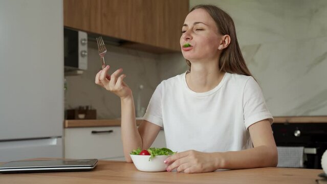 Young unsatisfied woman eats a salad sitting at home at the kitchen table. 