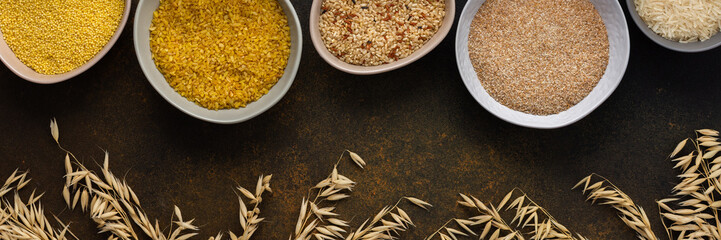 Various grain cereals in bowls banner, top view on a brown background with bowls of cereals and ears of oats