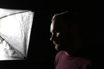 Portrait of a middle aged caucasian man's profile in shadow against a black background with studio light from softbox on his face. He is wearing a burgundy sweater.