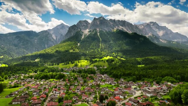 Aerial timelapse of majestic mountains of the Alps, with a small town in the valley and the clouds in the blue sky casting moving shadows

