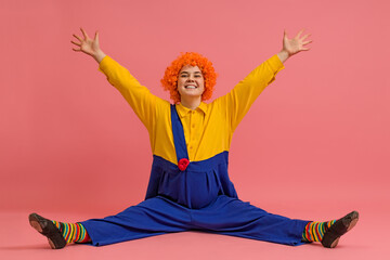 a joyful clown in a wig and a yellow-blue suit sits with his legs apart and raised his hands up on...