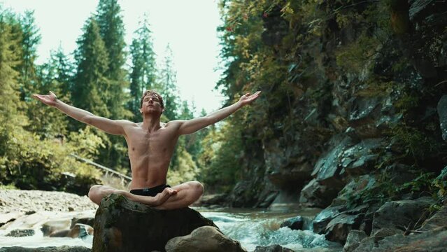 Confident red haired man doing breathing exercise on a stone in the running river 