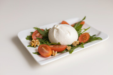 Fresh burrata cheese with arugula leaves, cherry tomatoes and walnuts on a white serving plate on a white table. Traditional Italian cheese