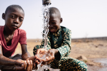 Two African toddlers playing with the water that flows from a rural faucet on the outskirts of their village; concept of water scarcity - obrazy, fototapety, plakaty
