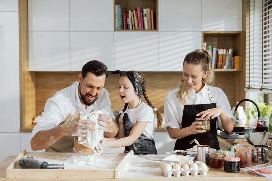 Curious daughter holding sive above wooden surface happy father pouring flour on it. Beautiful blonde mother preparing food for homemade pizza. Family cooking at weekend having fun.