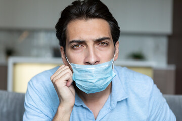 Close up portrait of sad man wearing medical mask in a wrong way and sitting on the couch and...