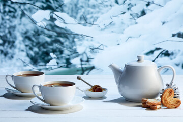 Obraz na płótnie Canvas Kettle and two cups of delicious black tea with honey and biscuits on winter forest background