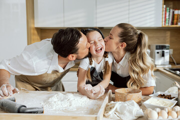 Pretty family in light kitchen. Father and mother kissing little daughter wearing aprons. Parents teaching daughter cooking kneading dough sieving flour preparing homemade pizza baking pasta cookies.