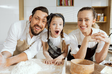 Curious daghter showing surprising emotions looking at camera. Happy father and mother looking laughing spending time together in modern kitchen at weekend.
