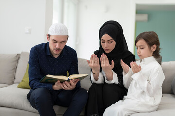 Traditional muslim family parents with children reading Quran and praying together on the sofa...