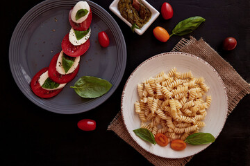  Italian pasta fusilli, caprese salad with tomato, basil mozzarella with pesto on black rustic background. Top view food. Italian dinner