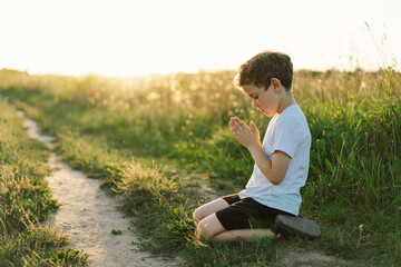 Boy closed her eyes and praying in a field at sunset. Hands folded in prayer concept for faith,...