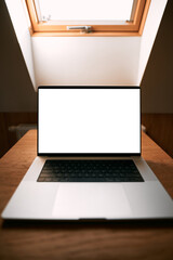 Opened modern professional laptop on a wooden table against opened roof window. Concept of working remotely in a cottage. Design mock-up with blank computer screen.