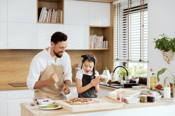 Delighted kid with father in aprons clapping in ahnds looking at selfmade pizza on wooden table. Preparing to put pizza in oven. Delighted parent having fun with beautiful daughter with braids.