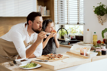 Curious daughter biting piece of homemade pizza standing at table in modern kitchen with father...