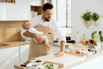 Smiling father holding baby in arms rolling dough with hands kneading on wooden surface cooking baking preparing domestic pizza.